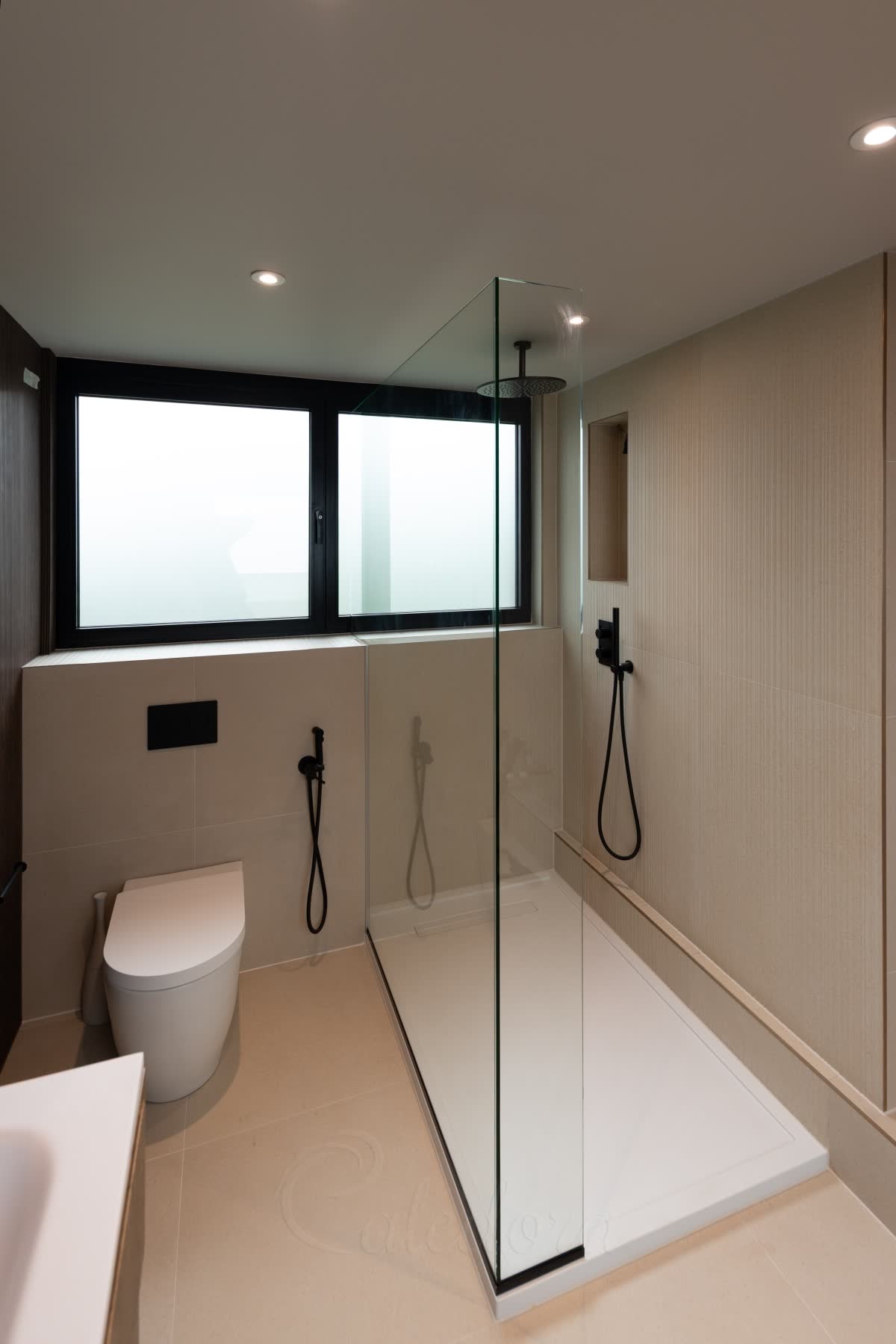 An L-shaped walk-in shower screen on a white shower tray — a tall clear glass panel running along the tray from the window wall, a short return joined to it at the outer corner, and a slim black profile at the foot of the glass turning the corner with it, with a black ceiling rain head and black controls beside a recessed niche
