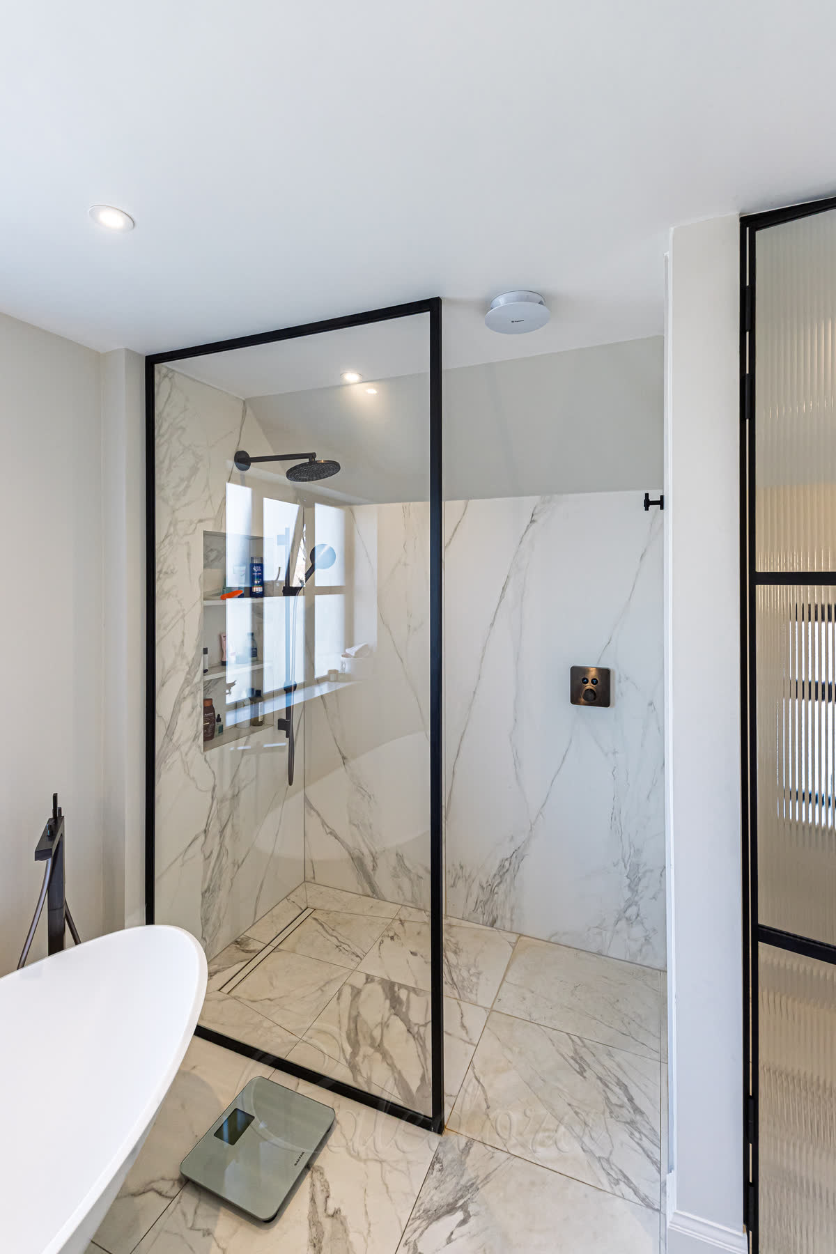 A walk-in panel in a slim matt black frame on a level marble-effect wet-room floor with a linear drain, a black rain head and hand shower inside and a freestanding bath beside it
