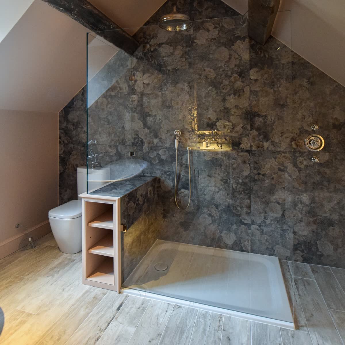 A walk-in screen standing on the edge of a white shower tray on a wood-effect floor, the tray plainly a separate piece from the floor around it, in a loft bathroom with dark floral tiles and a ceiling rain head