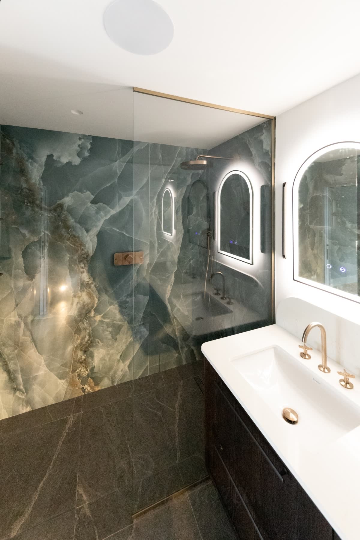 A single-panel walk-in shower screen — one clear glass panel coming off the wall and running up to the ceiling in a slim brass-finish channel, the entry beside it left open onto a level dark stone floor with a linear drain, blue onyx-effect walls inside the shower