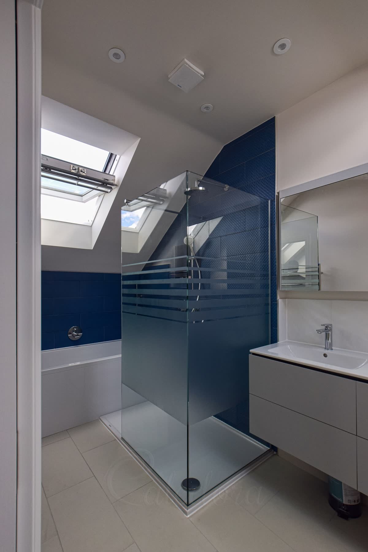 Two clear glass panels meeting at a glass-to-glass corner on a white shower tray, a band of frosted stripes across the middle, under a sloping loft ceiling with a roof window and deep blue tiles