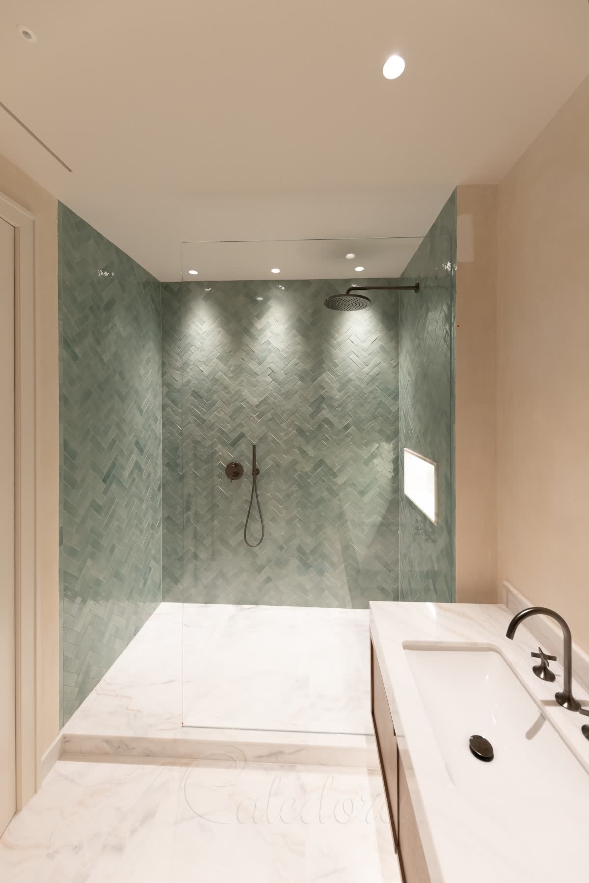 A clear walk-in panel standing on a raised white marble shower floor one step above the bathroom, sage green herringbone zellige tiles inside, a lit niche and gunmetal shower fittings