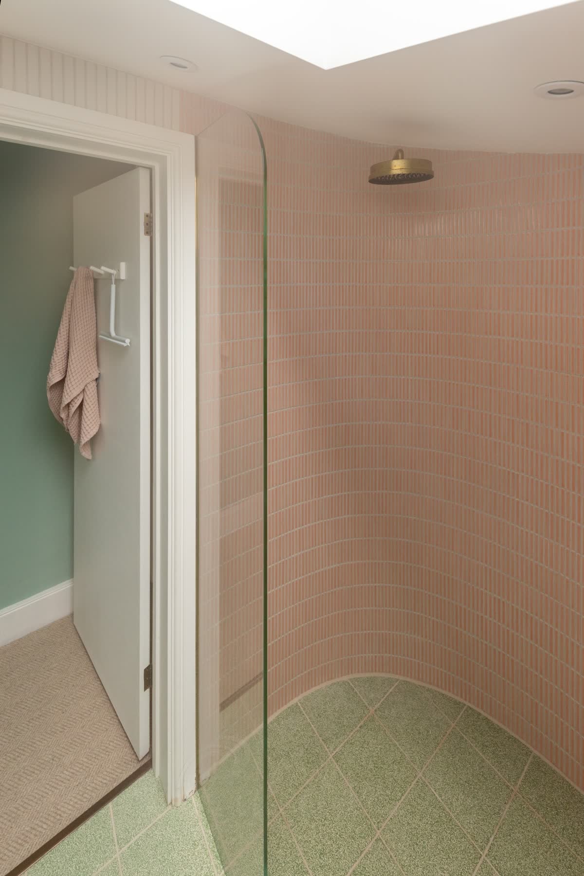 A clear walk-in panel with its outer top corner rounded off, standing on a level green terrazzo wet-room floor beside the doorway, the pink stick-tiled wall behind it curving round the shower under an aged brass rain head