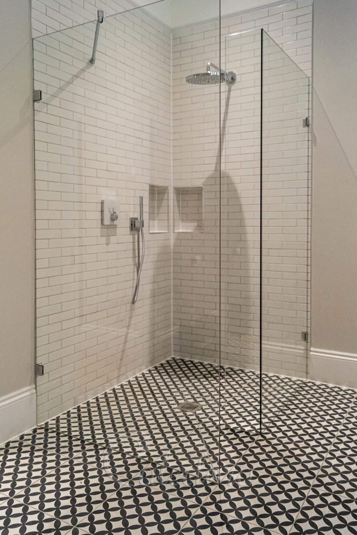 A traditional walk-in shower enclosure in a corner between two walls of white metro tiles — an L-shaped screen of two bonded glass panels off one wall, a second panel off the other wall held by an angled support bar, and the gap between them left open as the way in, on a level black and white patterned tile floor with a centre drain and a round rain head