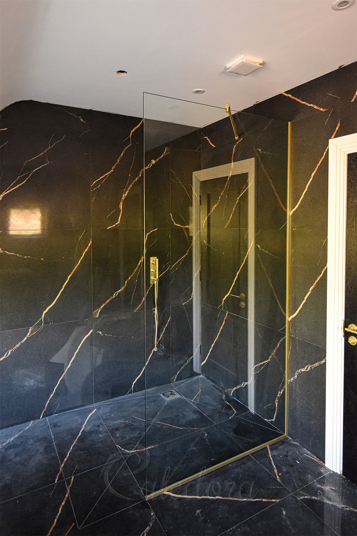 A clear walk-in panel steadied by a short brushed brass bar running back at 45° from its top edge to the wall, the glass held in brushed brass channels at the wall and floor, against black marble-effect tiles with gold veining