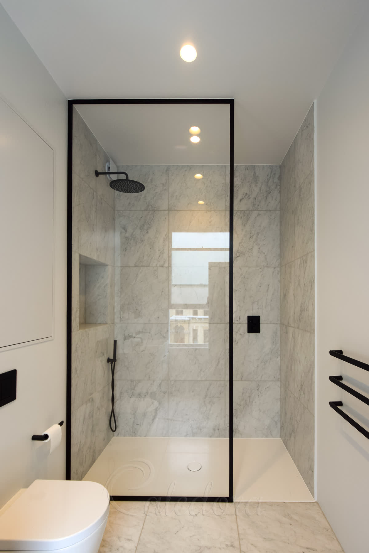 A black walk-in shower screen — one full-height clear panel inside a slim black frame, standing on a white shower tray against grey-veined marble walls, with a matching black rain head, controls and towel rail