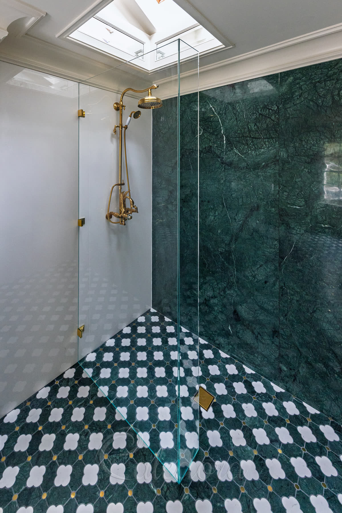 An L-shaped walk-in shower screen in ultra clear glass — the main panel held off a white wall by three polished brass brackets, a narrow return UV-bonded to it at the corner with no bracket of its own, standing on a green and white patterned marble wet-room floor beside a green marble wall, under a skylight