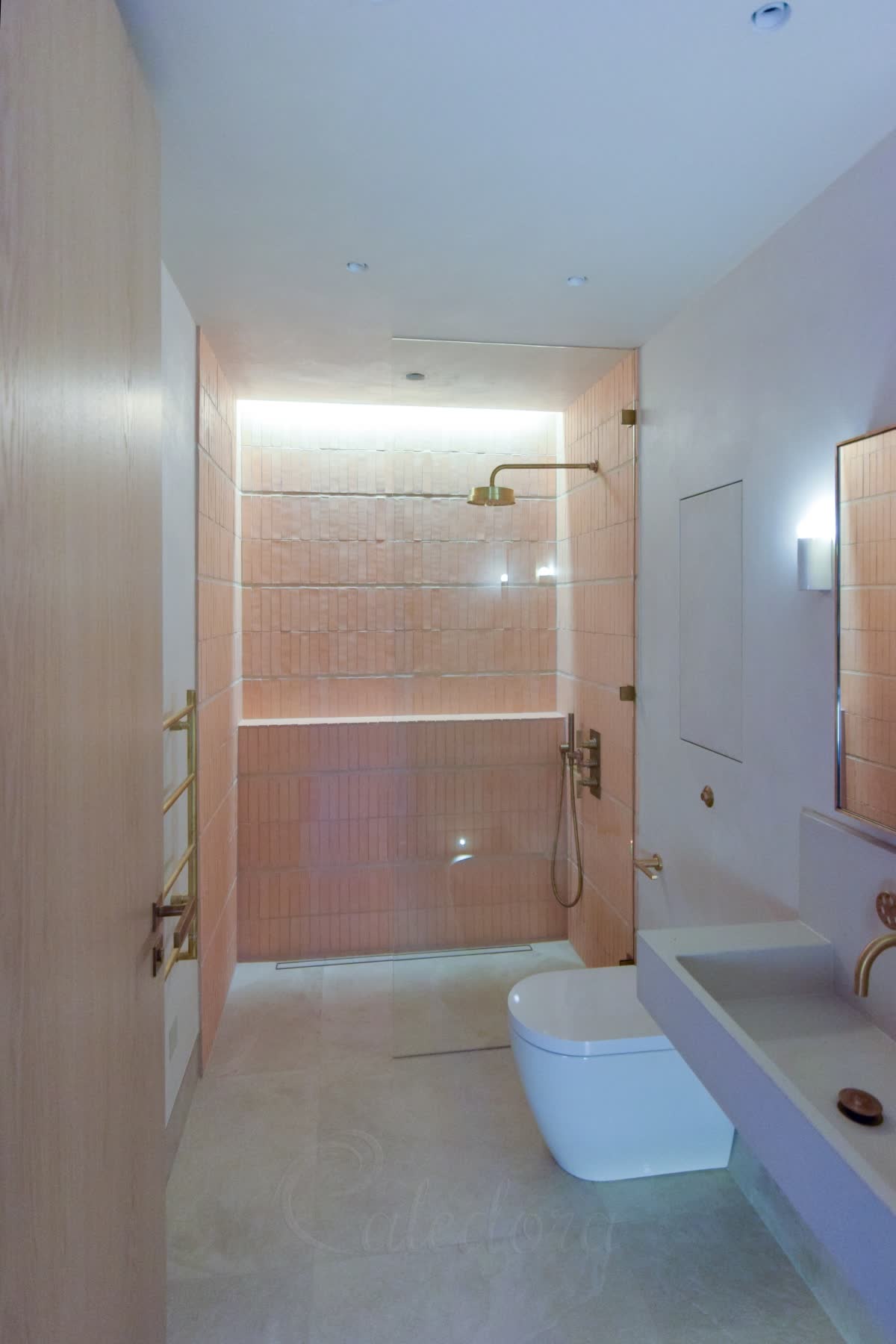 A clear walk-in panel held off the wall on small brushed brass clamps, the open side left as the way in, on a level stone wet-room floor with a linear drain, pink stacked tiles inside with a lit ledge and brushed brass shower fittings