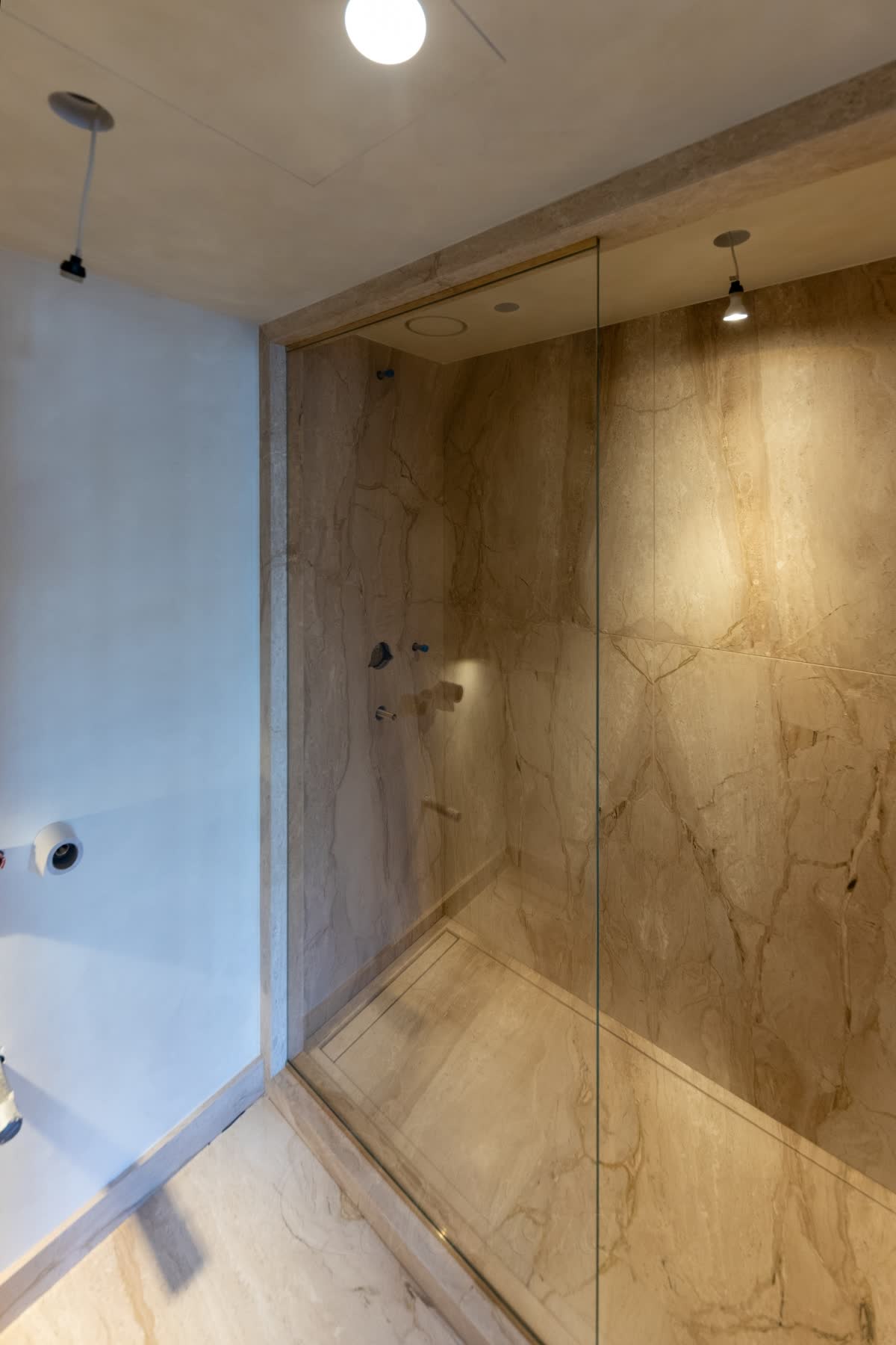 A clear walk-in panel in a slim brushed brass channel along the stone bulkhead above, the open end left as the way in, with the shower floor laid in the same beige marble-effect stone and a linear drain against the wall