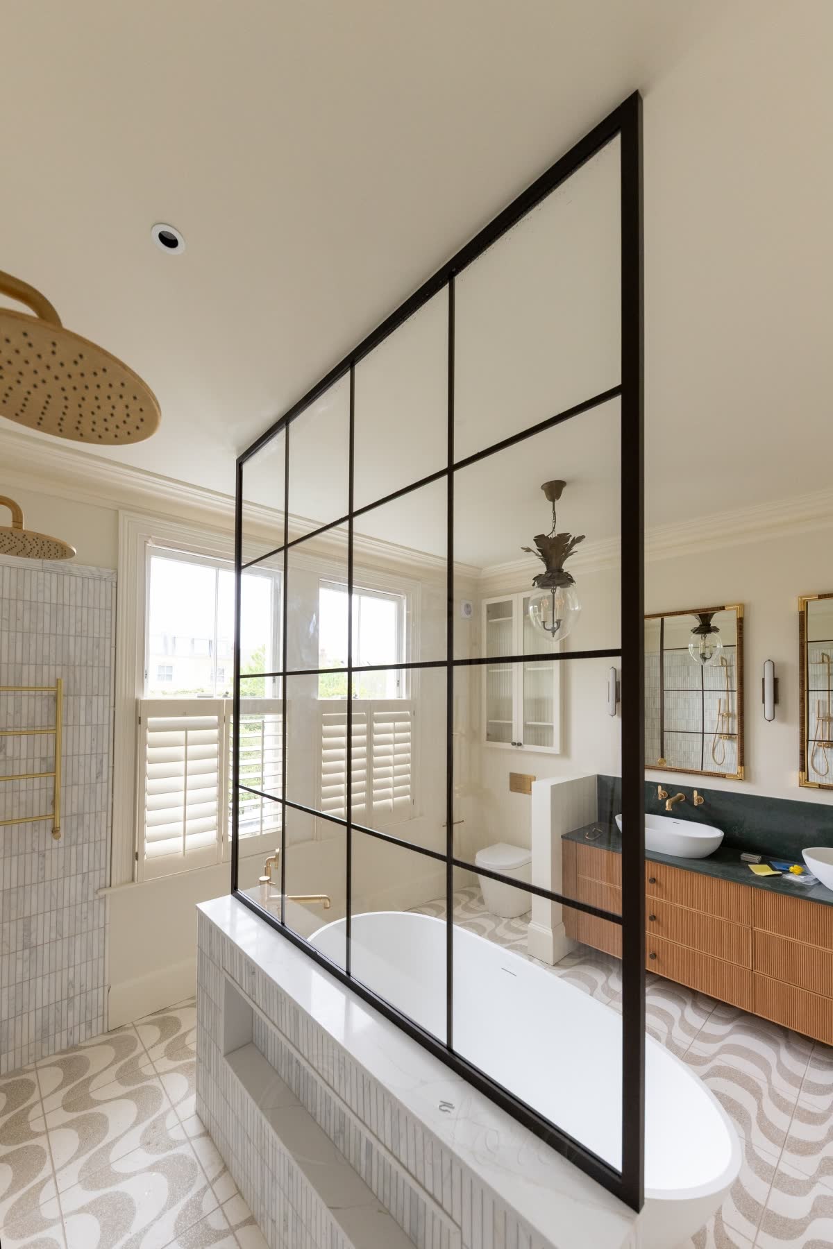 A dark bronze grid walk-in screen seen from inside the shower, standing on a tiled half-wall with a niche and rising to the ceiling, free-standing with no side wall, a freestanding bath beyond it and brushed brass rain heads overhead