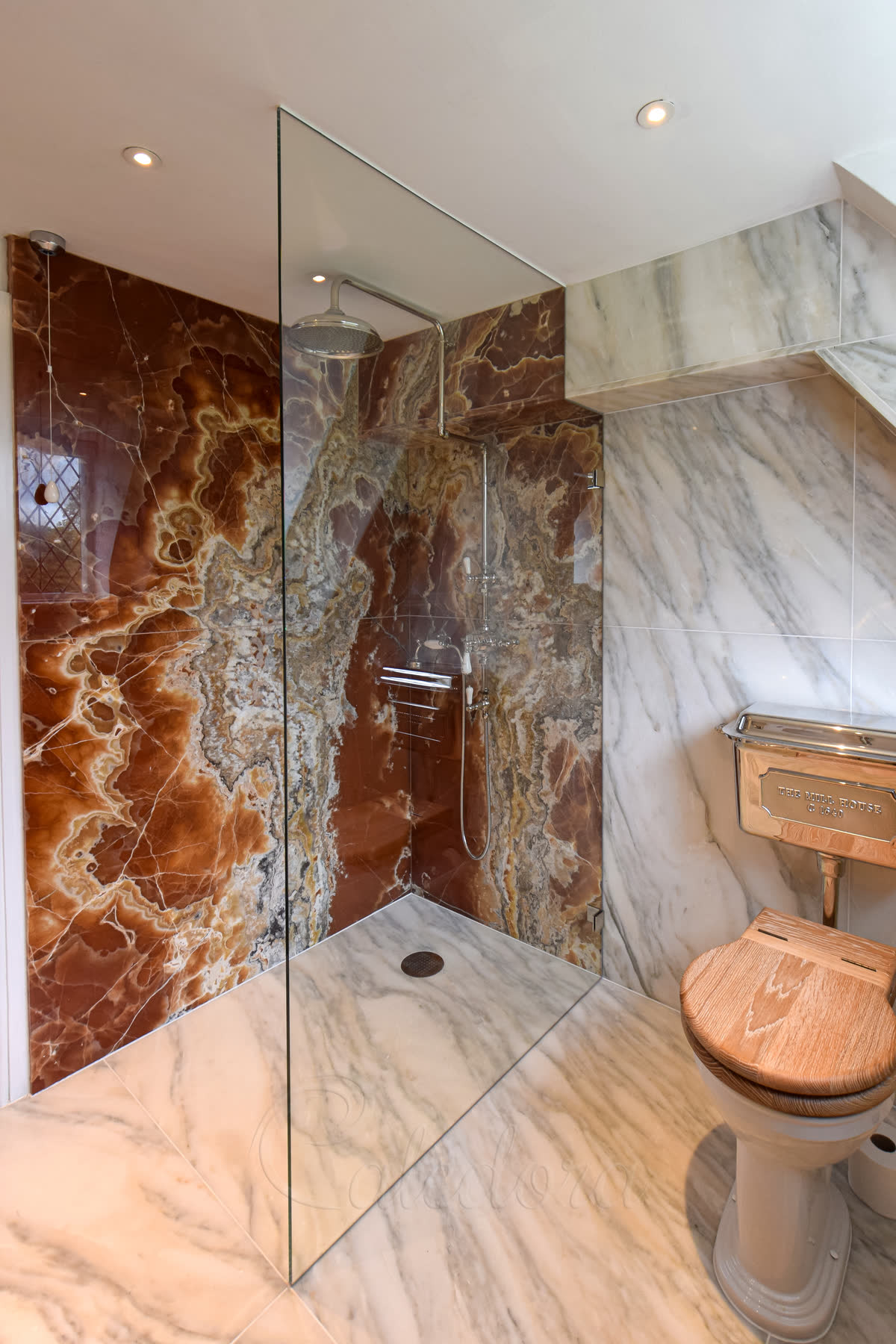 A clear walk-in panel notched at the top to fit round a marble-clad ceiling step, held on small polished clips, on a level white marble wet-room floor against walls of red and amber onyx