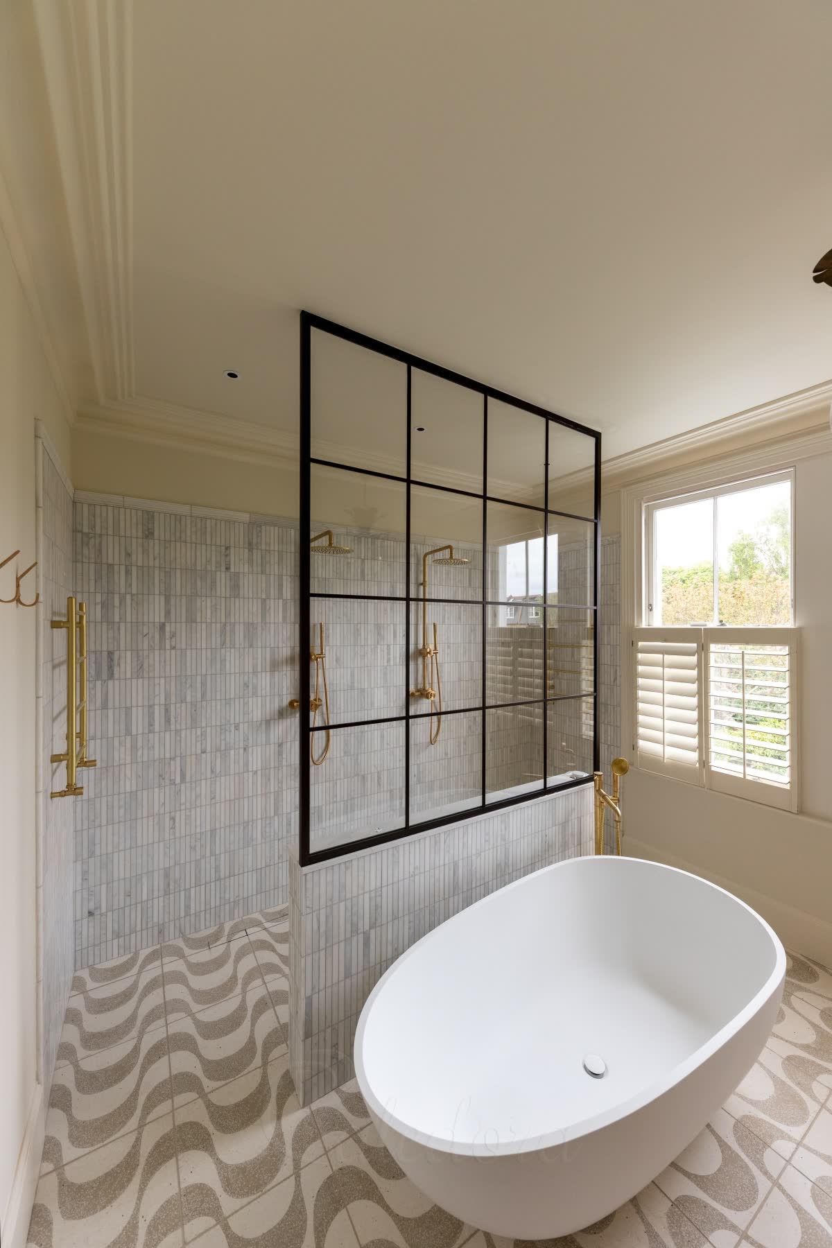 A dark bronze grid walk-in screen standing on a tiled half-wall and fixed to the ceiling, with no side wall holding it, the wave-patterned floor running past its open end into a shower with two brushed brass rain heads, and a freestanding bath in front of the wall