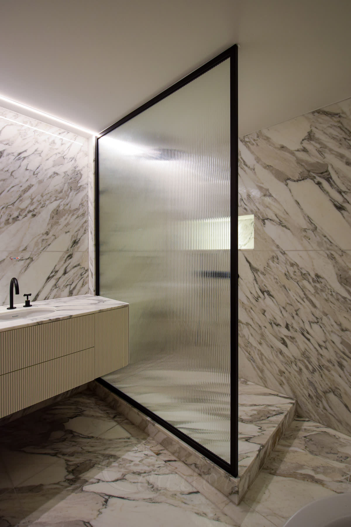 A raised wet-room floor 100 mm above the bathroom, finished in the same grey-veined white marble as the floor and walls, with a black-framed reeded glass walk-in panel standing on the raised floor and running up to the ceiling, beside a fluted vanity