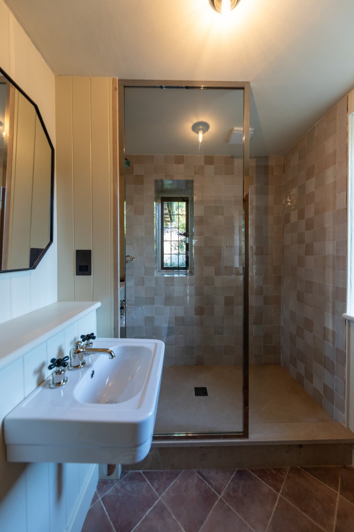 A polished nickel walk-in shower screen — one tall clear panel edged in slim nickel profiles, standing on a raised stone shower floor with blush zellige tiles inside and a leaded window, beside a basin with nickel taps