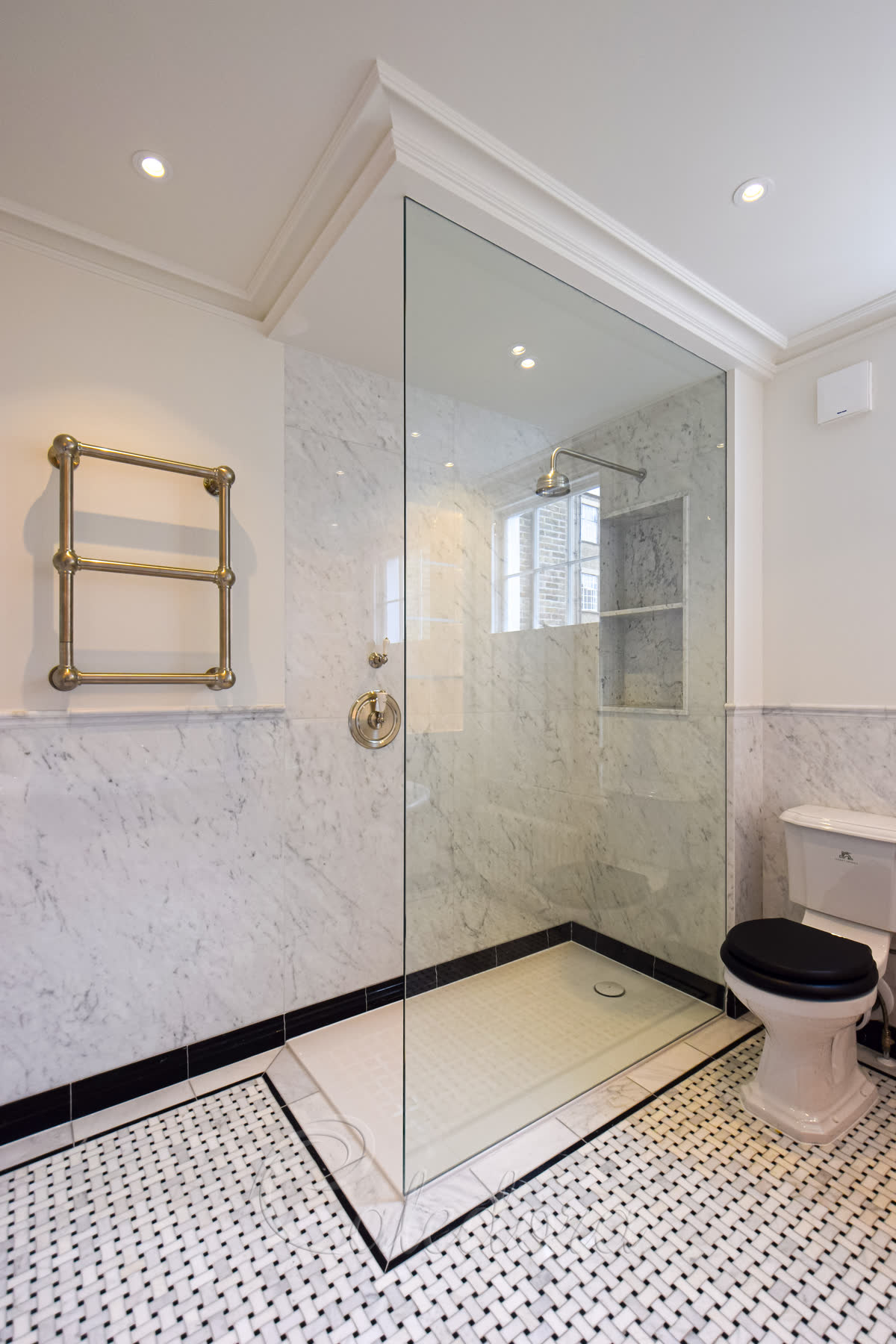 A clear walk-in panel set into the wall and running up into the cornice with no metal in sight, standing on a white tray inside a black-bordered basketweave marble floor, with white marble walls and a shelved niche