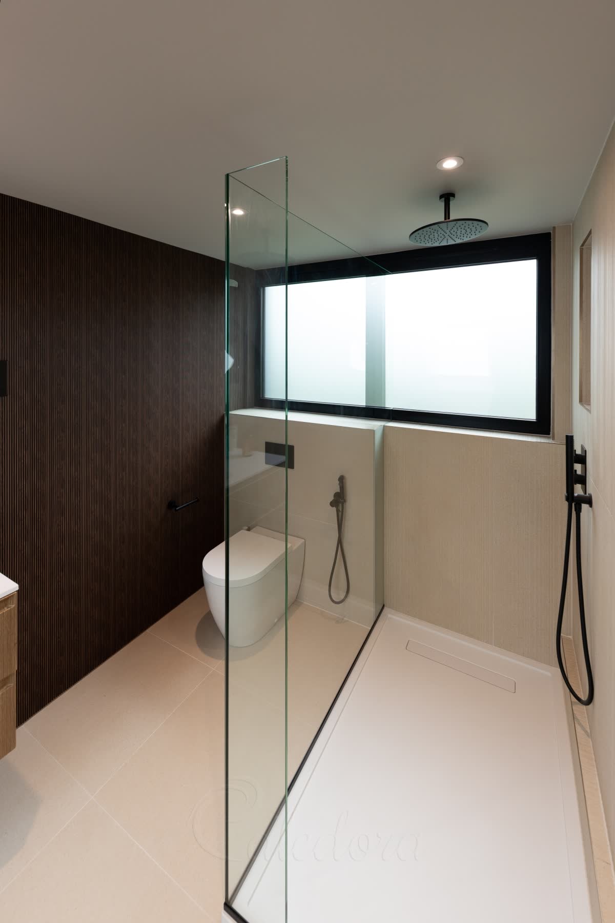 A walk-in screen seen from the shower entry — a clear glass panel along a white shower tray with a short return joined to it at the open end, the two forming an L that stands with no bar above and a clear gap below the ceiling, a slim black profile at the foot and a black rain head over the tray