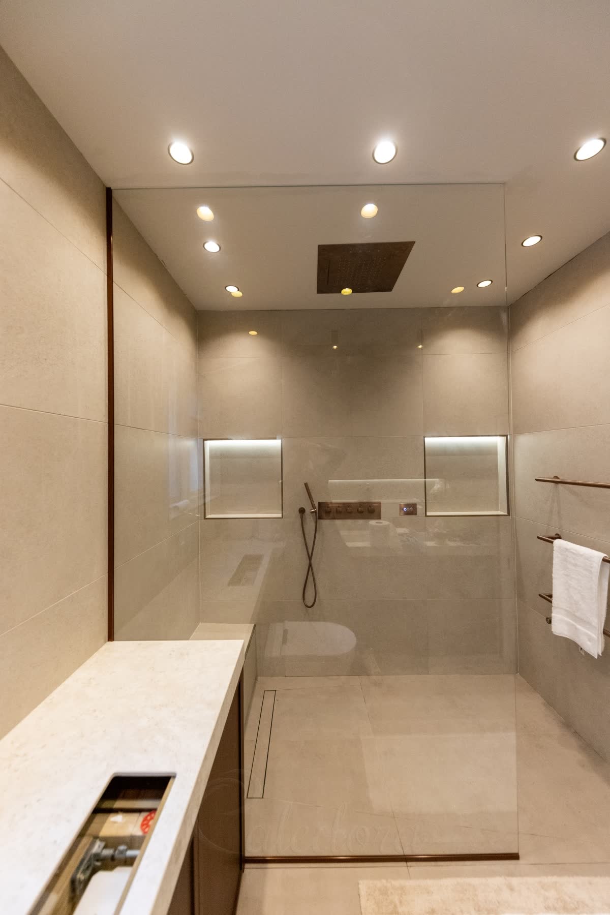 A clear walk-in panel rising to the ceiling in slim bronze-coloured channels along the wall and floor, a large ceiling rain head, two lit niches and a linear drain in a level stone-effect wet room