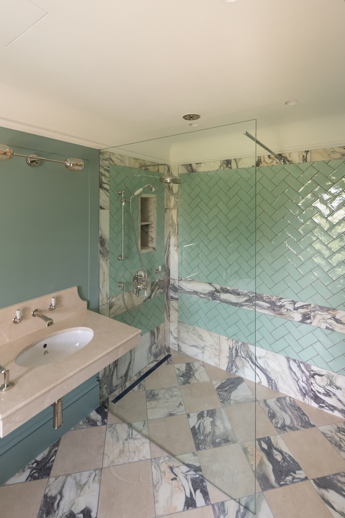 A clear walk-in panel stopping short of the ceiling, steadied by a slim polished bar running square from its open top corner to the wall, with green herringbone tiles and bands of veined marble behind, on a level chequered marble floor with a linear drain
