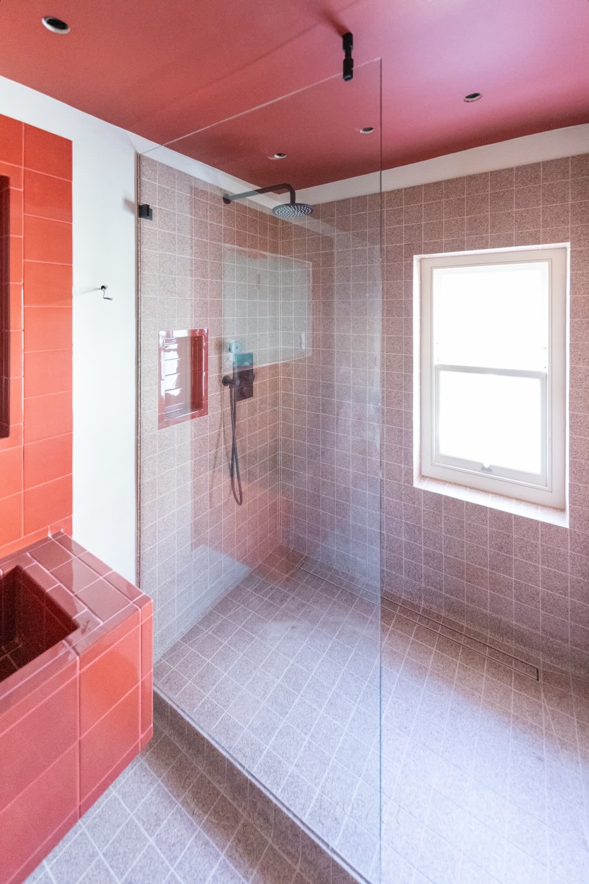 A clear walk-in panel held off a tiled wall by a small black clip, with a short black tube running from the top of its open edge straight up to a red ceiling across the gap above the glass, over a raised tiled shower floor with a linear drain