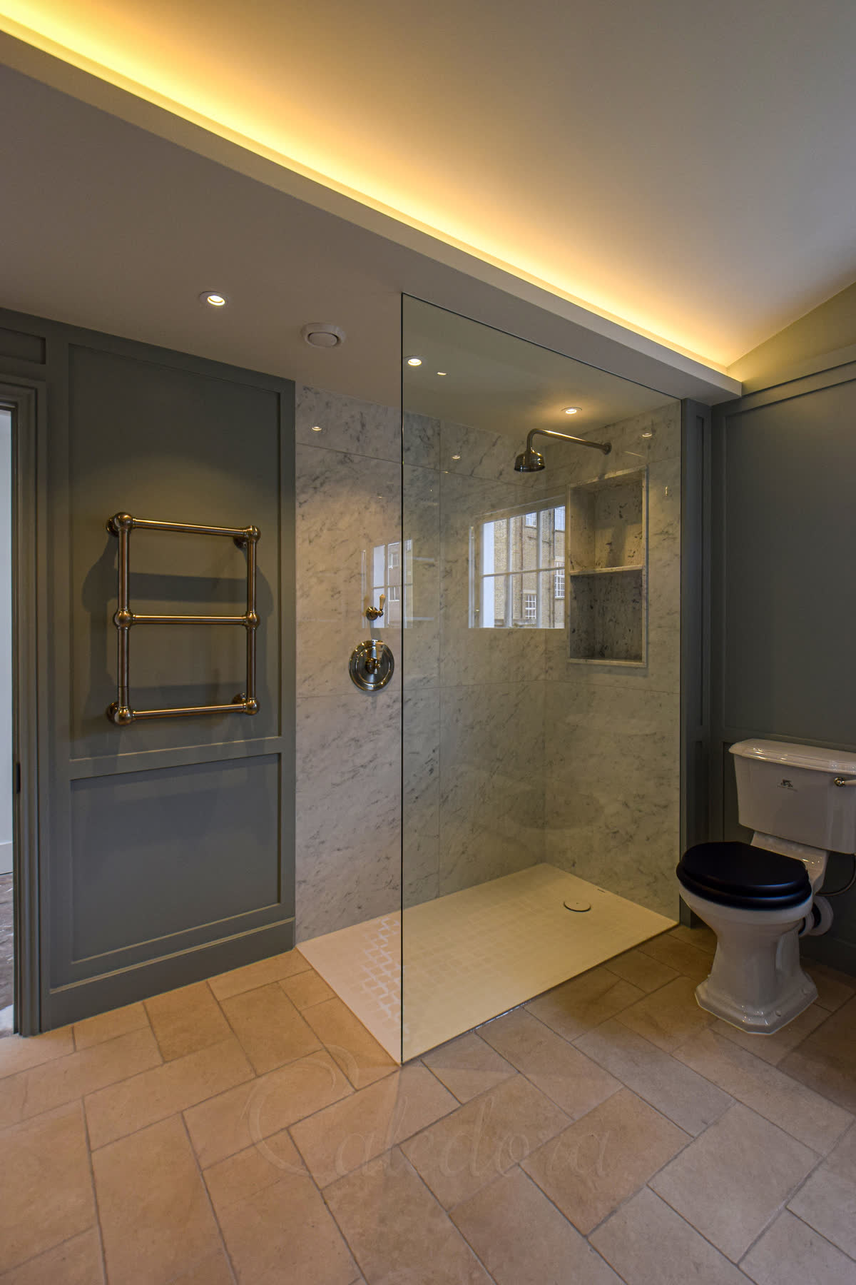 A clear walk-in panel with no visible metal rising to the ceiling from a wide white shower tray, white marble walls with a shelved niche and a window inside, nickel fittings, grey panelled walls and a herringbone limestone floor