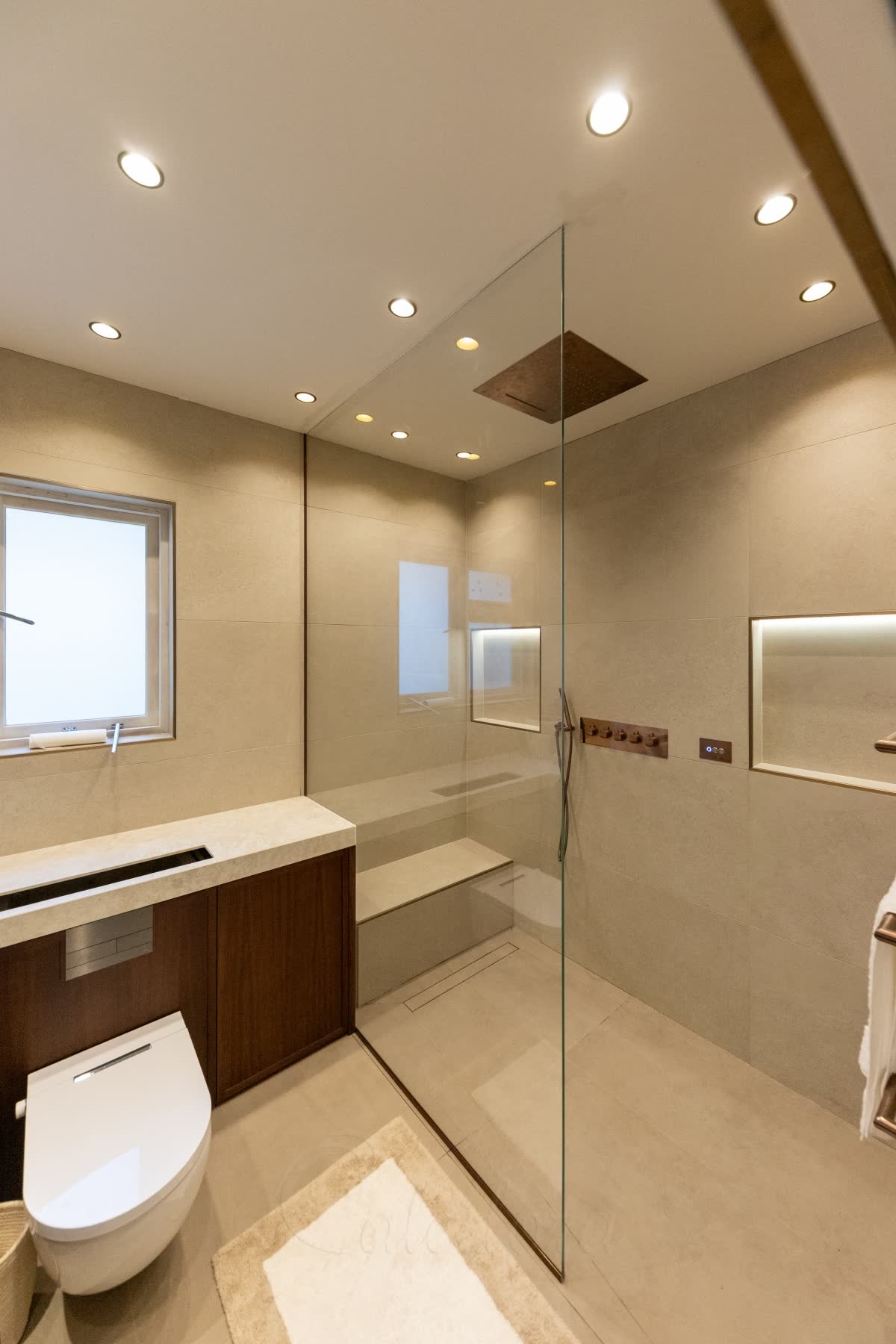 A walk-in shower with a bench rising from the floor — a solid stone-look tiled seat with a step, built against the back wall, behind a single full-height glass panel that runs up to the ceiling, with a linear drain at the foot of the bench, a bronze rain head and lit niches on a level wet-room floor