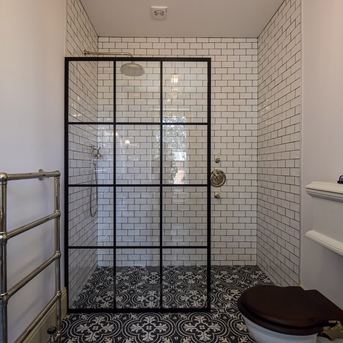 A black grid walk-in screen on a tiled wet-room floor, the same black and white patterned tiles running from the bathroom under the glass to the back wall of the shower with no visible tray, white metro tiles on the walls
