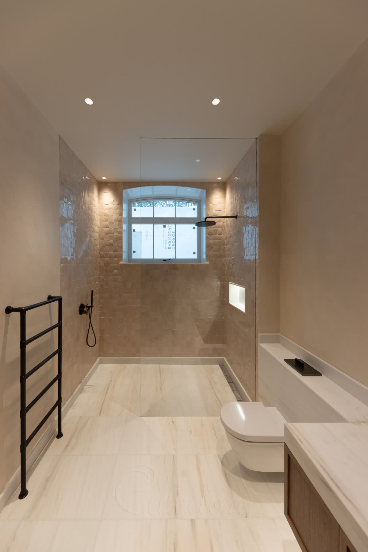 A clear walk-in panel with no visible metal on a level white marble wet-room floor with a linear drain, beige zellige tiles inside around a round-topped window, black shower fittings and a black towel rail
