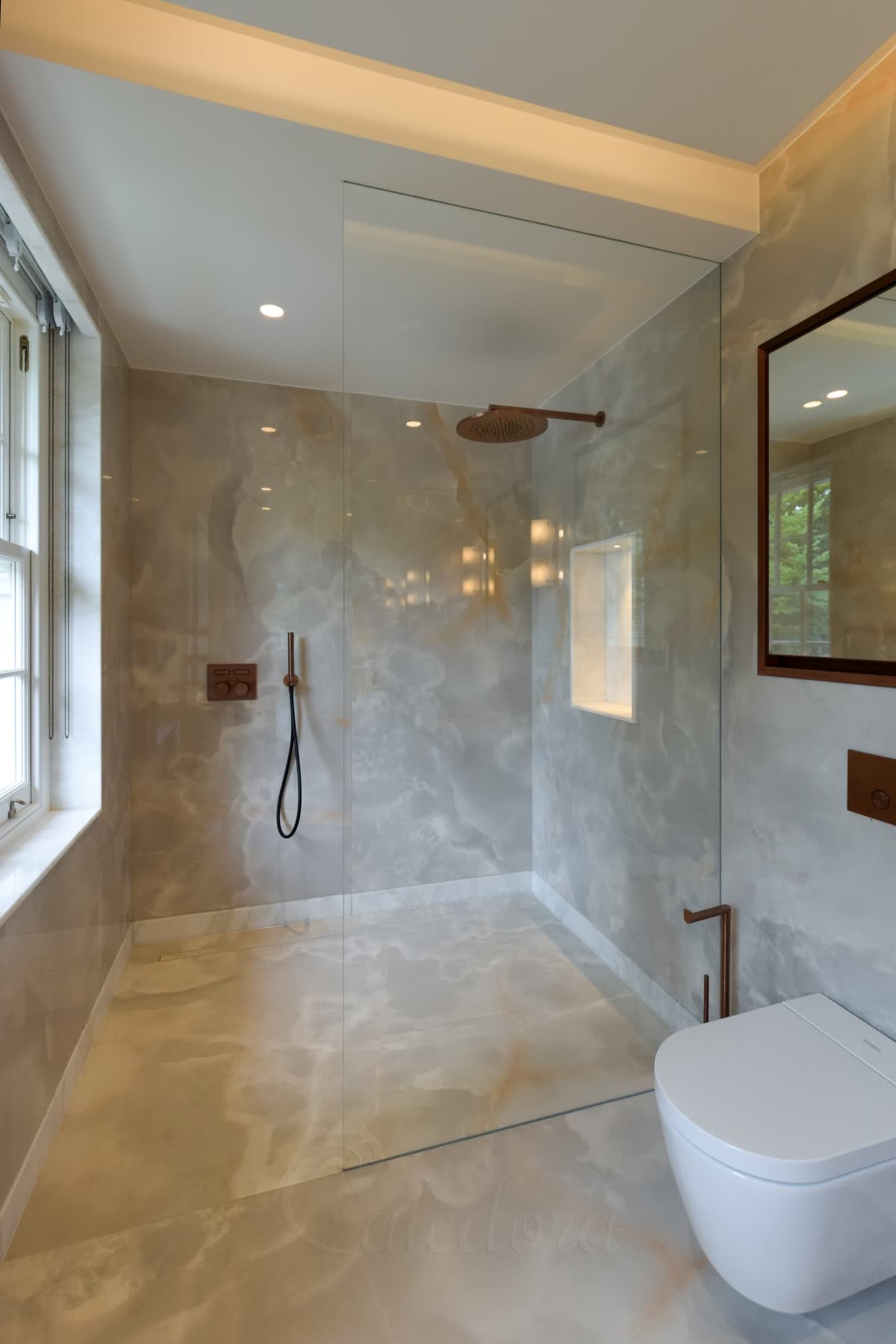A wet room shower screen with no metal anywhere — one clear glass panel set into grooves cut in the stone floor, the wall and the ceiling and sealed with mastic in the same colour, rising from a level onyx-patterned stone floor that runs on unbroken from the bathroom to a linear drain