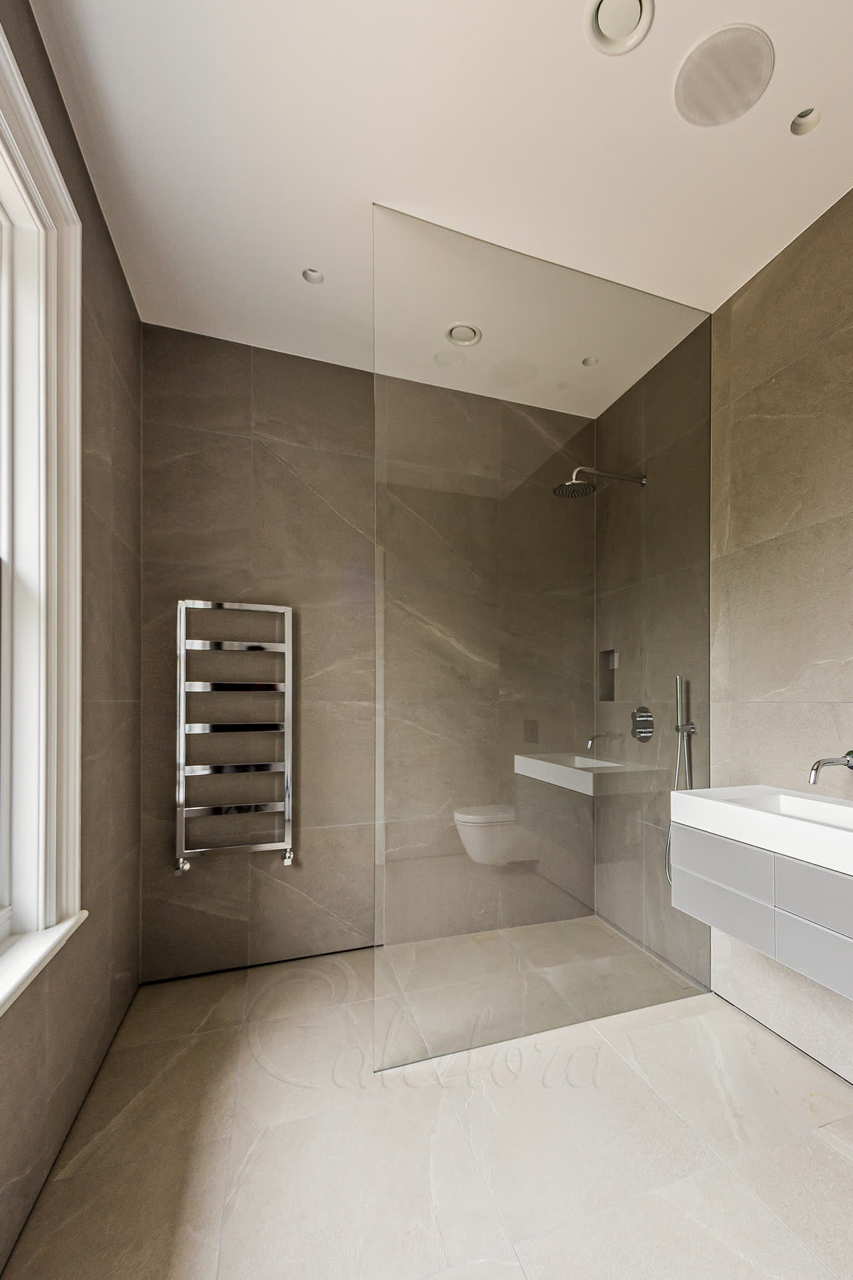 A wet room shower screen with no metal fixings — a single 10 mm ultra clear panel set into grooves in the wall and the ceiling and sealed with mastic matched to the tiles, over a level tiled floor that runs on from the bathroom with no step