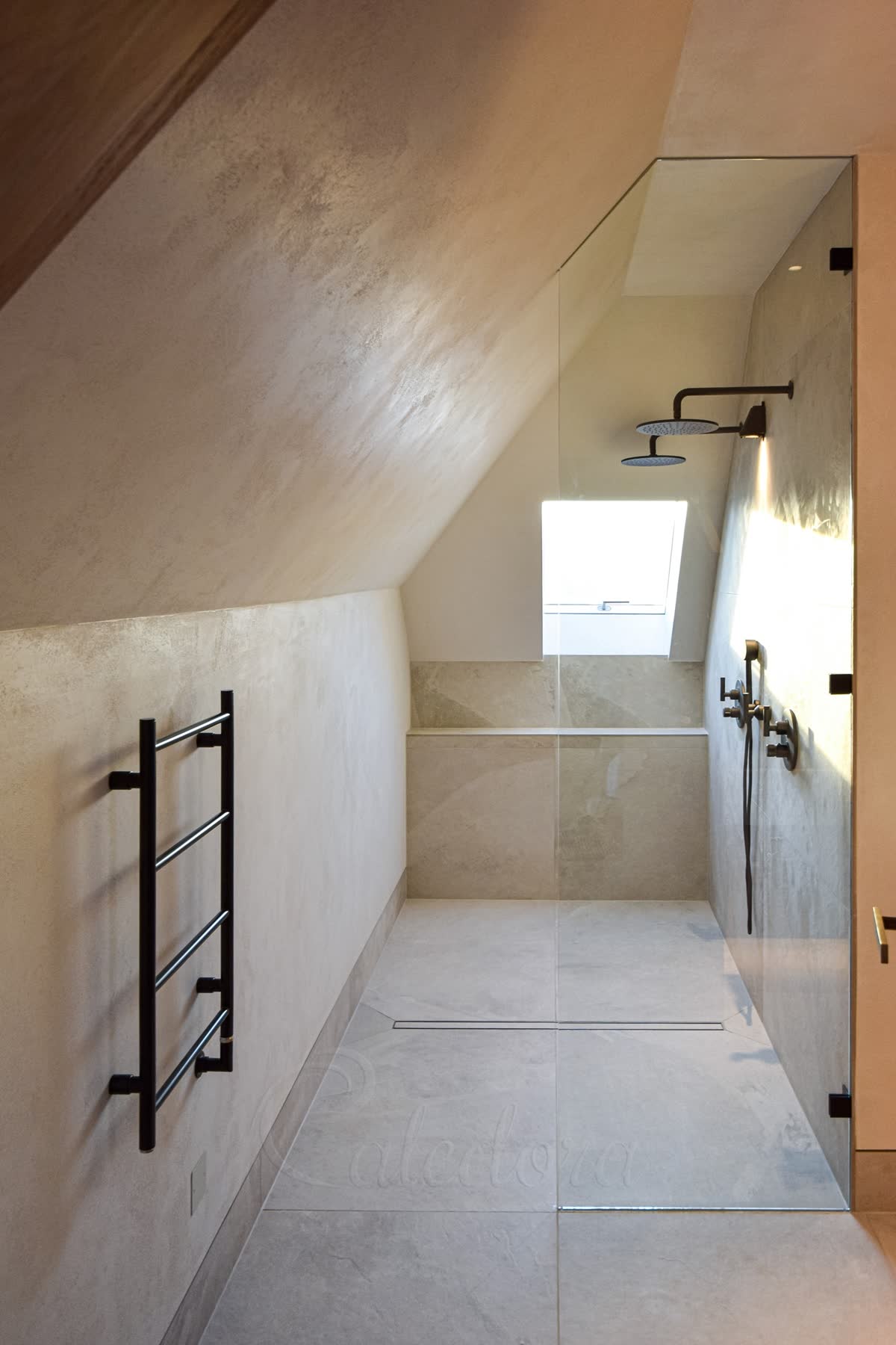 A tall clear walk-in panel on three black wall clamps, its top edge cut to follow the sloping ceiling, on a level stone-effect wet-room floor with a linear drain, in a long loft shower under a roof window with two black rain heads