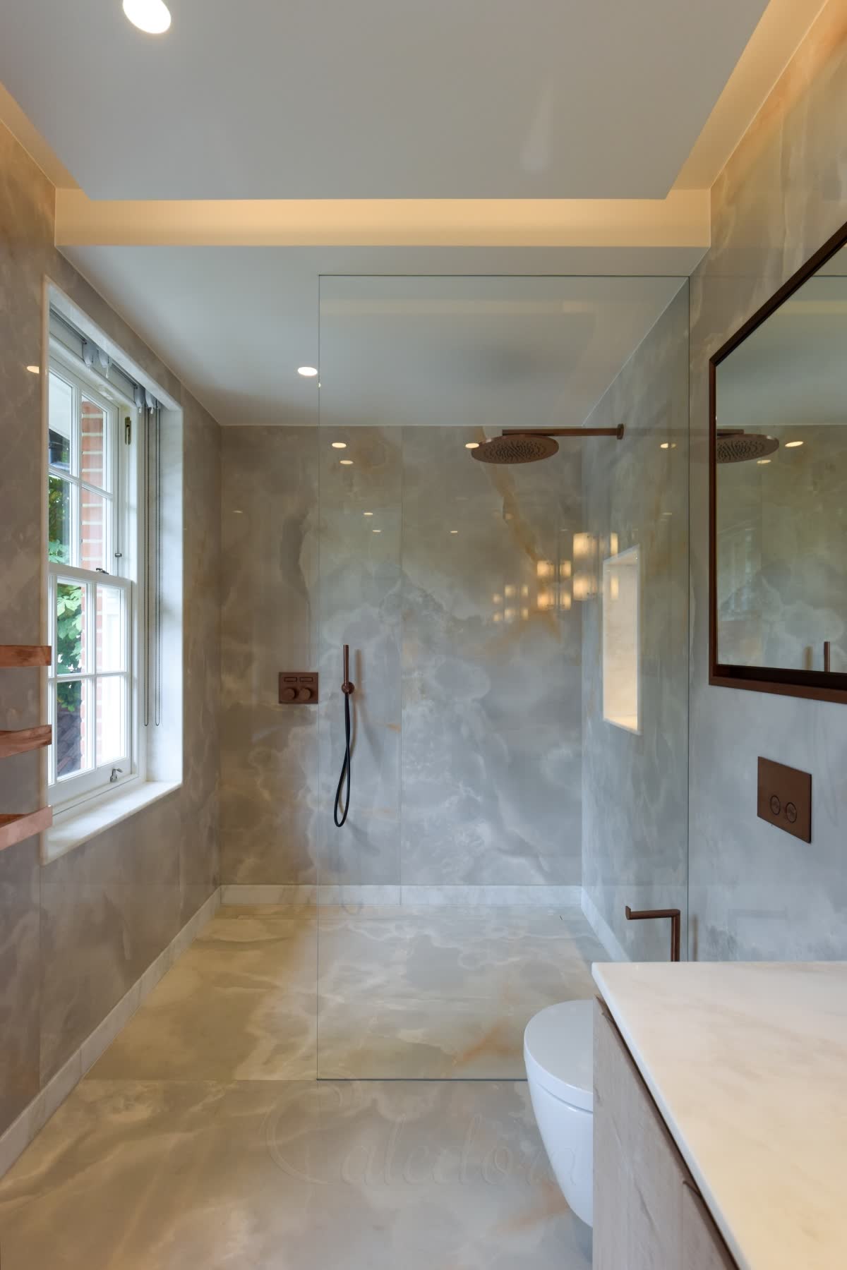 A clear walk-in panel with no visible metal, rising from a level onyx-effect wet-room floor into the lit ceiling recess, the same stone running on into the bathroom, with copper-coloured shower fittings and a sash window beside it
