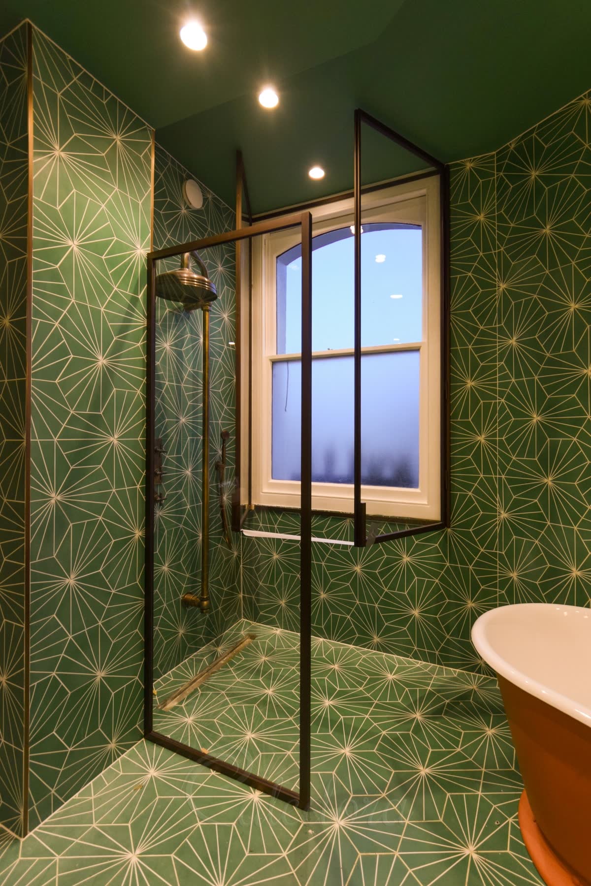 A walk-in screen in a slim dark frame on a tiled wet-room floor with a linear drain, a second framed panel set back round the sash window, green star-pattern tiles over the walls and floor and an aged brass exposed shower
